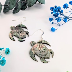Pode incluir: Um par de brincos prateados com design de tartaruga. Os brincos são iridescentes com um padrão esculpido na concha. A imagem é estilizada com raminhos de flores azuis e folhagem verde em um fundo branco.