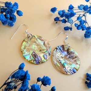 Könnte beinhalten: Ein Paar runde Ohrringe aus schillerndem Abalone-Muschelmaterial mit silberfarbenen Haken. Die Ohrringe zeigen eine Mischung aus Gold-, Grün- und Blautönen. Blaue Blumen sind auf beigem Hintergrund um die Ohrringe angeordnet.