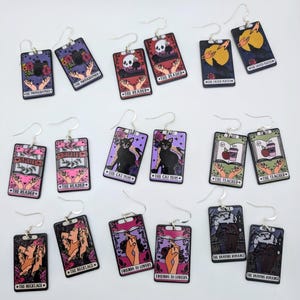 May include: A selection of rectangular earrings with silver hooks. Each earring displays a tarot card design with illustrations and text, such as "The Photographer," "The Reader," and "The Cat Mom." The colour scheme includes purple, pink, and black.