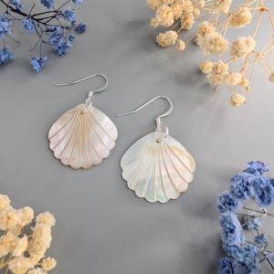 以下が含まれることがあります： 銀色のフックが付いた貝殻型のイヤリングのペア。イヤリングは真珠のような白で、扇のようなデザインです。背景はグレーで、隅にはドライフラワーが飾られています。