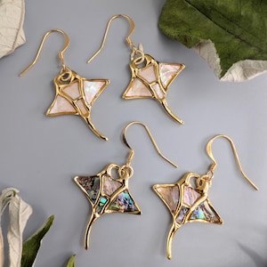 May include: Four pairs of gold-colored stingray earrings. The earrings have a gold outline with a mosaic-like inlay of white and iridescent shell pieces. The earrings are displayed on a gray surface with green leaves.