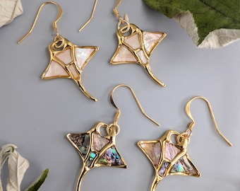 Puede incluir: Cuatro pares de pendientes dorados con forma de manta raya. Los pendientes tienen un contorno dorado con una incrustación de mosaico de piezas blancas e iridiscentes. Los pendientes se muestran sobre una superficie gris con hojas verdes.