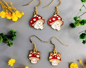 May include: Four gold-tone earrings. Two pairs feature red mushroom caps with white spots and smiling white creatures. The other two earrings have red mushroom caps with white spots and cat faces. Yellow and green floral accents surround the earrings.