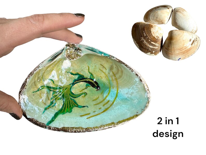Puede incluir: Un plato de concha de mar con un pez verde y negro pintado en su interior. La concha es de color azul claro y tiene un borde dorado. El texto "2 in 1 design" es visible debajo de la concha.