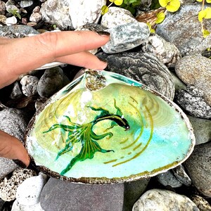 Puede incluir: Una concha de mar con un pez pintado de verde y negro en su interior. La concha est&aacute; sentada sobre un lecho de rocas.