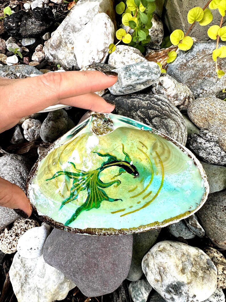 Puede incluir: Una concha de mar con un pez pintado de verde y negro en su interior. La concha est&aacute; sentada sobre un lecho de rocas.