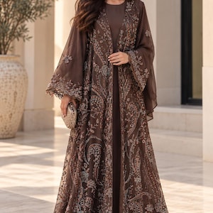Könnte beinhalten: Ein bodenlanges braunes Kleid kombiniert mit einem passenden, vorne offenen, transparenten Mantel. Der Mantel ist mit aufwendigen Stickereien, Perlen und Pailletten in Blumen- und Paisleymustern verziert und hat gewellte Säume an den weiten Ärmeln.