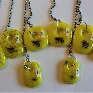 May include: Eight yellow glass pendants with a honeycomb pattern and a bee design. Each pendant has a silver chain and a number from 1 to 8.