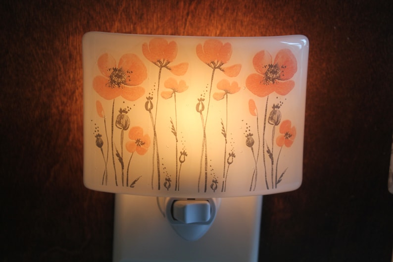 Może przedstawiać: Biała ceramiczna lampka nocna z kwiatowym wzorem. Wz&oacute;r przedstawia pomarańczowe kwiaty maku z szarymi łodygami i liśćmi. Lampka nocna ma biały włącznik/wyłącznik.