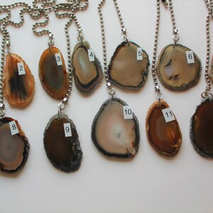 May include: A collection of twelve agate slice pendants, each with a unique pattern of brown, tan, and cream colors. The pendants are strung on silver ball chains, ideal for jewelry making or craft projects.