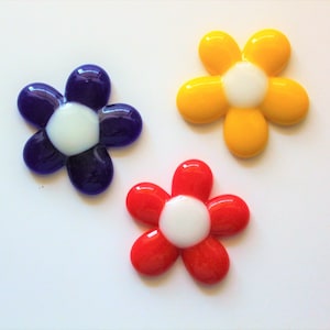 May include: Three colorful glass flower-shaped ornaments with white centers. The flowers are purple, yellow, and red.