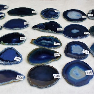 May include: A collection of 16 blue agate slices, each with a unique pattern and shape. The slices are arranged in rows on a white surface. The numbers 94 through 114 are written on the slices.