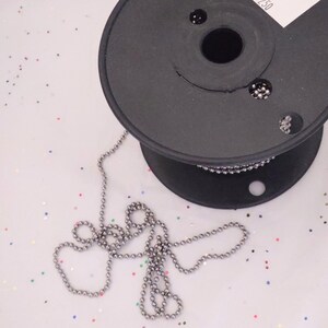 May include: A spool of silver chain with a black plastic spool. The chain is made of small, round beads.
