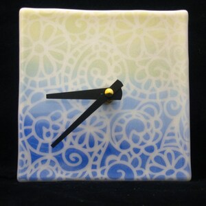 May include: A square ceramic clock with a white background and a blue and yellow ombre design. The clock face has a white background with a black hour and minute hand and a gold knob.