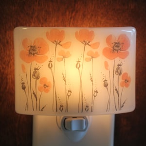 Może przedstawiać: Biała ceramiczna lampka nocna z kwiatowym wzorem przedstawiającym pomarańczowe maki i szare łodygi. Lampka nocna jest podłączona do gniazdka i ma biały przełącznik.
