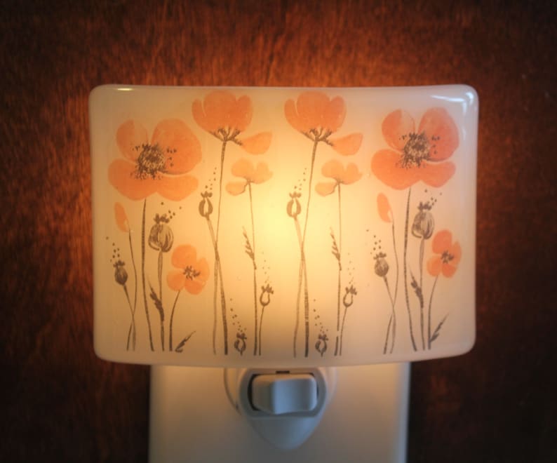 Może przedstawiać: Biała ceramiczna lampka nocna z kwiatowym wzorem przedstawiającym pomarańczowe maki i szare łodygi. Lampka nocna jest podłączona do gniazdka i ma biały przełącznik.