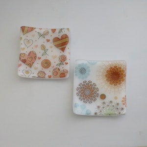 May include: Two square glass dishes with decorative patterns. One dish has a white background with brown and pink hearts and flowers. The other dish has a white background with brown and blue floral designs.