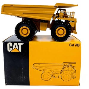 May include: A yellow Caterpillar 789 mining dump truck scale model with black tires and detailed cab features. The truck sits atop its original rectangular cardboard packaging, which is yellow and black with the CAT logo and Cat 789 text printed.