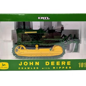 May include: A green and yellow John Deere 1010 crawler with ripper toy model inside a green ERTL box. The packaging features the text John Deere 1010 Crawler with Ripper and the John Deere logo. The model includes detailed tracks and a rear ripper.