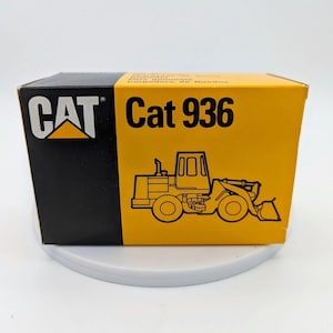 May include: A rectangular cardboard box with a yellow and black color scheme. The left side features the CAT logo in white and yellow on a black background. The right side displays the text Cat 936 above a black line drawing of a wheel loader.