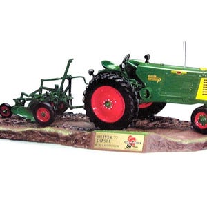 May include: A green and yellow Oliver 77 Diesel tractor model with red wheels, pulling a matching green plow. The tractor features a vertical exhaust pipe, a seat, and a plaque on the base reading Oliver 77 Diesel with 50 years branding.
