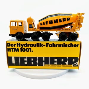 May include: A yellow toy concrete mixer truck with the words "LIEBHERR" on the side. The truck is on a yellow box with the words "Der Hydraulik-Fahrmischer HTM 1001. LIEBHERR So baut man Hydraulik-Fahrmischer." printed on it.