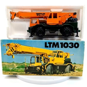 May include: An orange scale model crane with black wheels and the text TREFFLER printed on the side, resting in a white styrofoam insert. Below it is a blue cardboard box featuring a yellow crane illustration and the text LTM 1030.