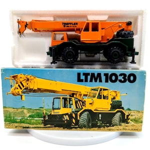May include: A yellow and black toy crane with the text "LTM 1030" on the box. The crane is on a dirt road with a green background. The box is white with the text "TREFFLER" on it.