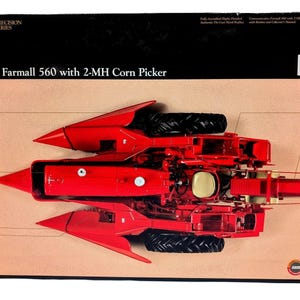 May include: A red die-cast metal replica of a Farmall 560 tractor with a 2-MH corn picker attachment. The packaging features the text 'The Farmall 560 with 2-MH Corn Picker' and 'Precision Series' against a black and tan background.