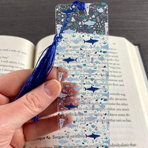 May include: A clear rectangular bookmark featuring a repeating pattern of various shark species, small fish, and seaweed in shades of blue and white. The bookmark has a hole at the top with a long, dark blue decorative tassel attached to it.
