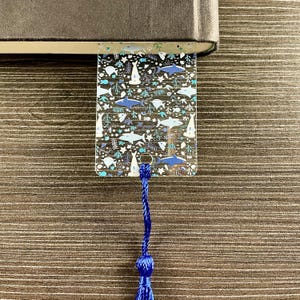 May include: A clear rectangular bookmark featuring a repeating pattern of blue and white sharks, fish, and seaweed. The bookmark has a small hole at the bottom with a dark blue braided cord tassel attached, and it is tucked into a dark book.
