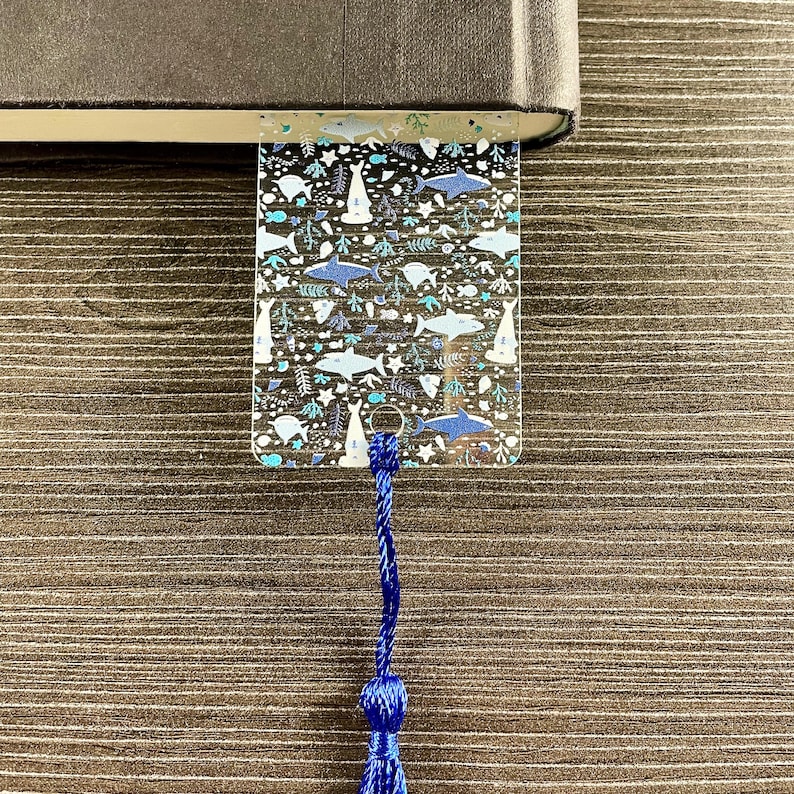 May include: A clear rectangular bookmark featuring a repeating pattern of blue and white sharks, fish, and seaweed. The bookmark has a small hole at the bottom with a dark blue braided cord tassel attached, and it is tucked into a dark book.