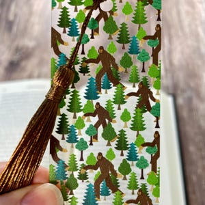 May include: A clear rectangular bookmark featuring a repeating pattern of brown Bigfoot figures and various green pine trees. The bookmark has a hole at the top with a long, dark brown decorative tassel attached to it.