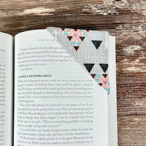 May include: A fabric bookmark with a geometric pattern of black triangles, coral triangles, and white arrows on a gray background. The bookmark is placed in a book with text visible. The bookmark is a useful accessory for readers.