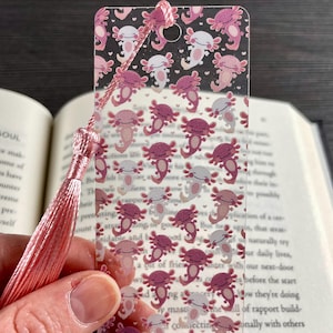 May include: A clear rectangular bookmark featuring a repeating pattern of small pink and white axolotl illustrations and tiny hearts. The bookmark has a hole at the top with a light pink silky tassel attached to it, resting against an open book.