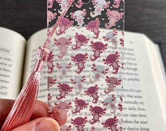 May include: A clear rectangular bookmark featuring a repeating pattern of small pink and white axolotl illustrations and tiny hearts. The bookmark has a hole at the top with a light pink silky tassel attached to it, resting against an open book.