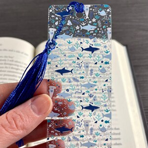 May include: A rectangular, transparent acrylic bookmark featuring a repeating pattern of various sharks, including hammerheads, in shades of dark blue, light blue, and teal, accented with small sea plants and stars, finished with a dark blue tassel.