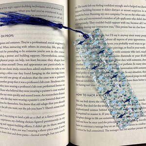 May include: A rectangular, translucent bookmark featuring a pattern of small, multi-colored shark silhouettes in shades of blue, grey, and white, accented with silver glitter. The bookmark is topped with a dark blue fabric tassel.