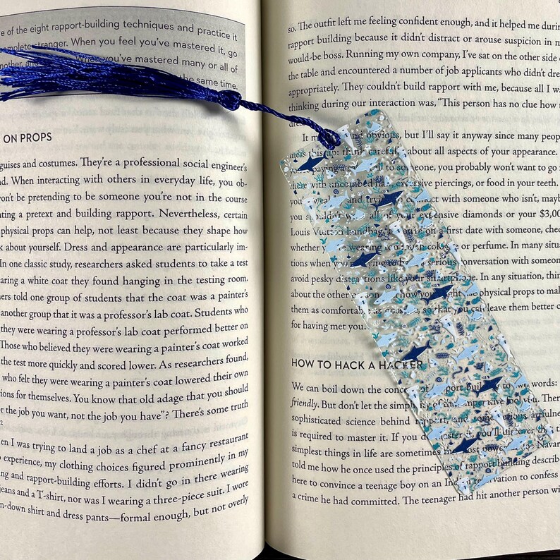 May include: A rectangular, translucent bookmark featuring a pattern of small, multi-colored shark silhouettes in shades of blue, grey, and white, accented with silver glitter. The bookmark is topped with a dark blue fabric tassel.