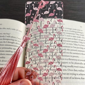 May include: A clear plastic bookmark featuring a pink flamingo pattern. The bookmark has a pink tassel attached to the top.