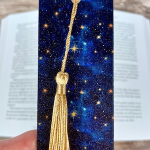 May include: A rectangular bookmark featuring a deep blue galaxy print with scattered yellow stars and nebulae. The bookmark includes a gold-colored cord attached to a matching gold-colored tassel that hangs from the top center of the item.
