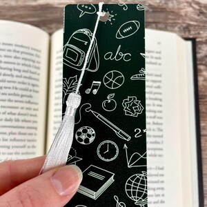 May include: A rectangular dark green bookmark featuring a white line-art pattern of school-related icons including a backpack, the letters abc, a football, a basketball, a pencil, a clock, a globe, and a book, finished with a white tassel.