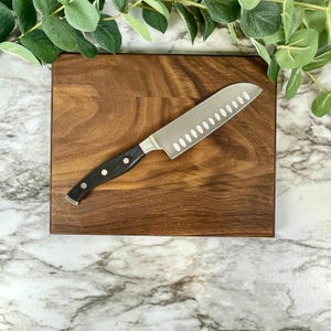 Puede incluir: Un cuchillo de acero inoxidable con mango negro descansa sobre una tabla de cortar rectangular de madera. El cuchillo tiene un borde dentado. La tabla de cortar es de color marrón intenso y tiene un patrón de vetas de madera natural.