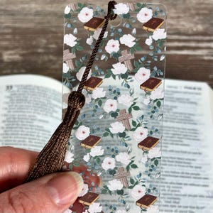 Peut inclure: Un marque-page rectangulaire en acrylique transparent orné d'un motif répétitif de fleurs blanches, de feuilles vertes, de croix en bois marron et de petits livres marron. Il possède un trou en haut avec un cordon tressé et un pompon marron.