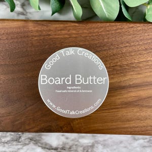 May include: A round, silver container of "Board Butter" sits on a wooden surface. The label reads "Good Talk Creations" and lists ingredients: food-safe mineral oil & beeswax. The website www.GoodTalkCreations.com is also printed on the label.
