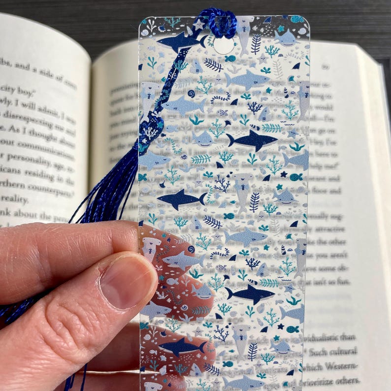 May include: A rectangular, semi-transparent bookmark featuring a repeating pattern of various sharks, including hammerheads, in shades of dark blue and light blue, interspersed with small teal coral and seaweed motifs, finished with a dark blue tassel.