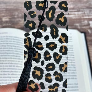 Puede incluir: Marcapáginas transparente con diseño de estampado de leopardo en negro y marrón. Tiene una borla negra. El marcapáginas está colocado en un libro abierto.