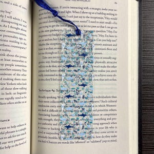 May include: A rectangular, clear acrylic bookmark featuring a pattern of small dark blue and light blue sharks, fish, and aquatic plants. The bookmark has a hole at the top with a dark blue tassel attached, resting vertically on an open book page.