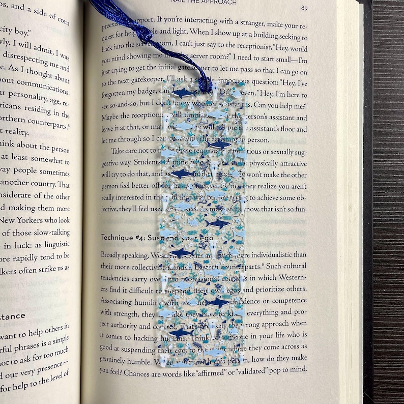May include: A rectangular, clear acrylic bookmark featuring a pattern of small dark blue and light blue sharks, fish, and aquatic plants. The bookmark has a hole at the top with a dark blue tassel attached, resting vertically on an open book page.
