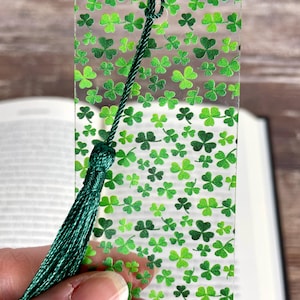 May include: A clear bookmark with a pattern of green shamrocks in varying shades. A dark green tassel hangs from the top. The bookmark is held in front of an open book.